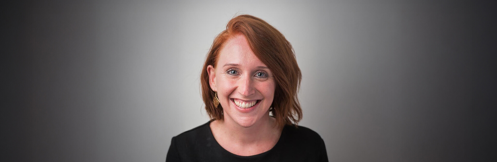 A smiling woman with short red hair and earrings, against a softly blurred gray background.