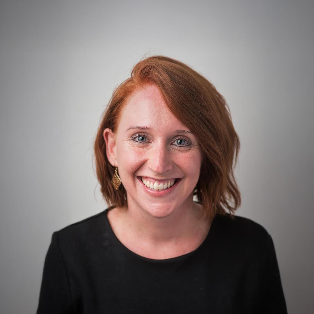 A smiling woman with short, wavy red hair, wearing a black top and earrings, against a light gray background.