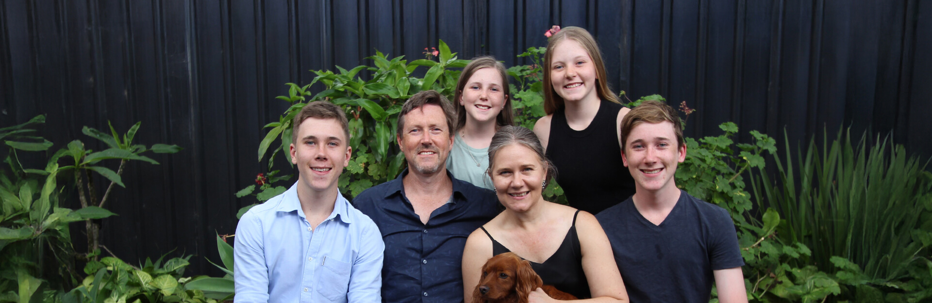A family of six, including two adults and four children, poses together outdoors with greenery in the background.
