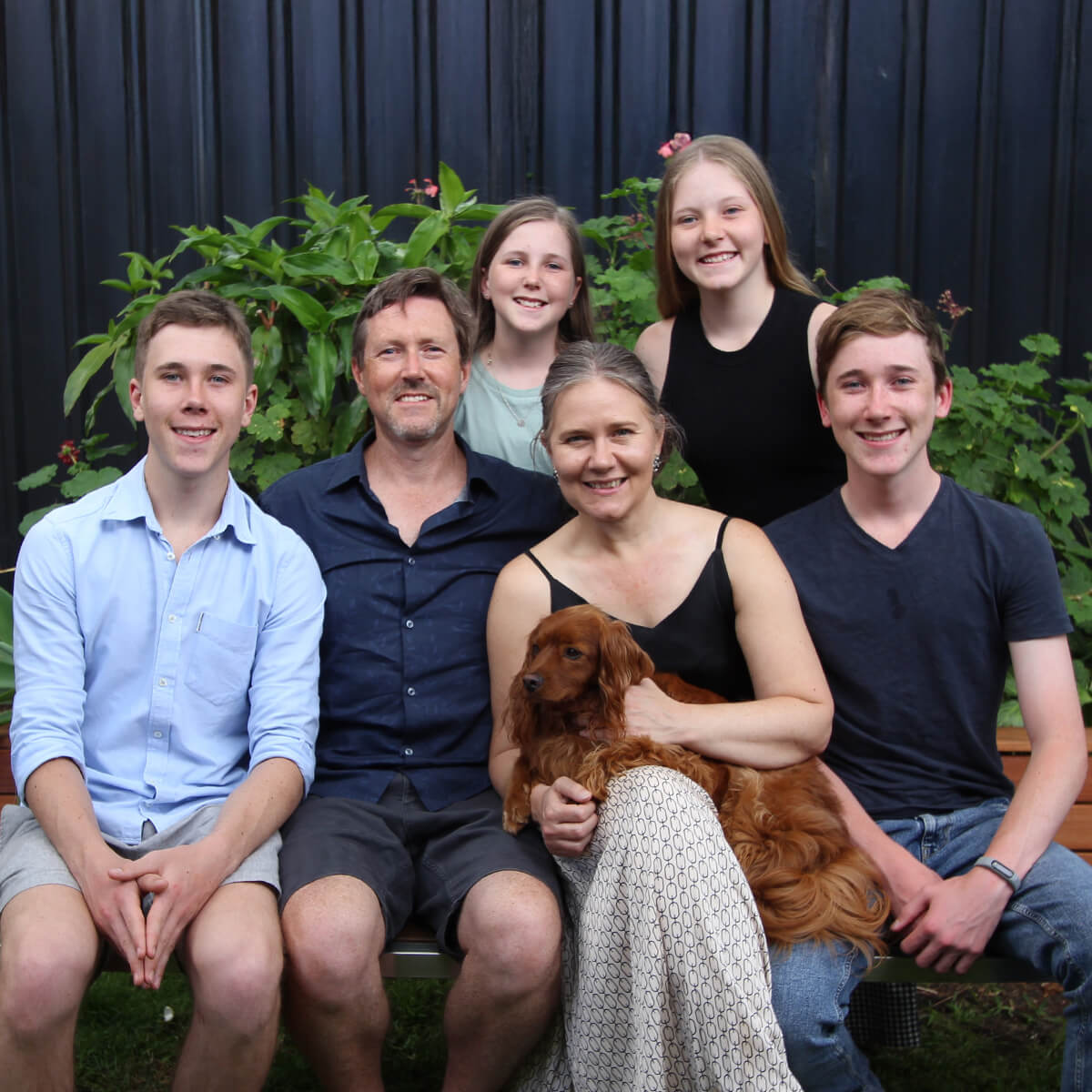 A family of six poses together outdoors, seated on a bench with a dog, surrounded by green plants.
