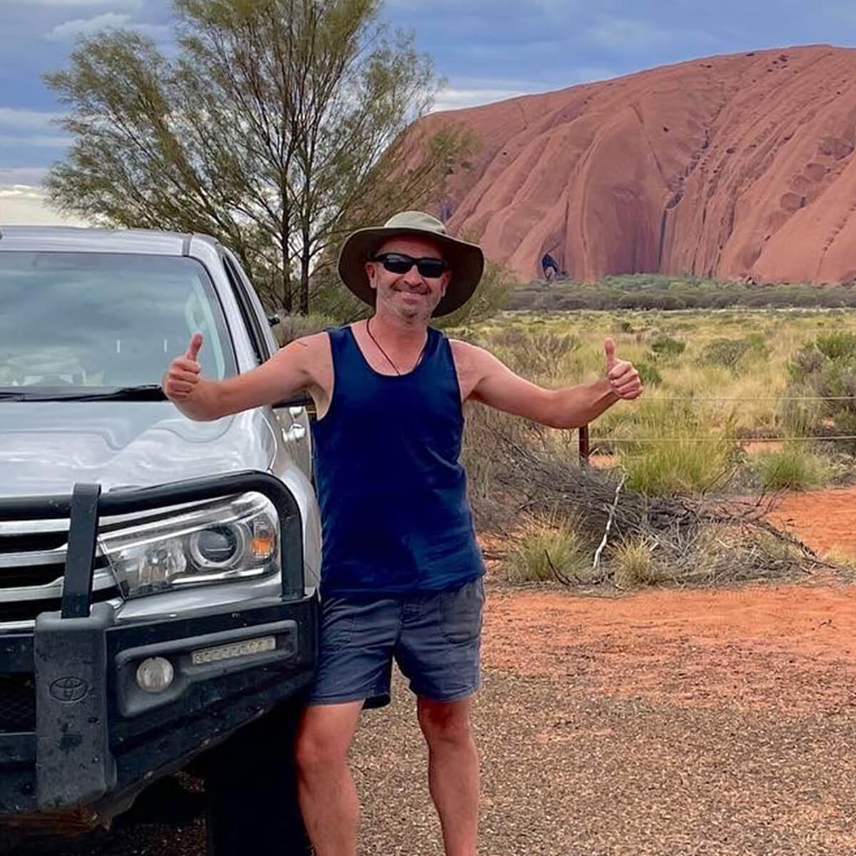 A man in a tank top and hat stands next to a vehicle, giving thumbs up with a red rock formation in the background.