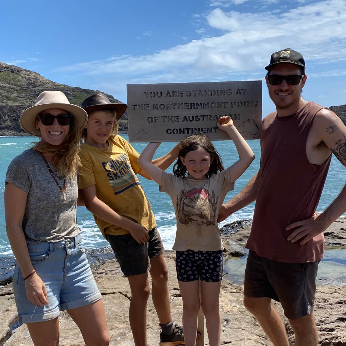 A family of four stands on rocky terrain by the sea, holding a sign that reads about the northernmost point of Australia.