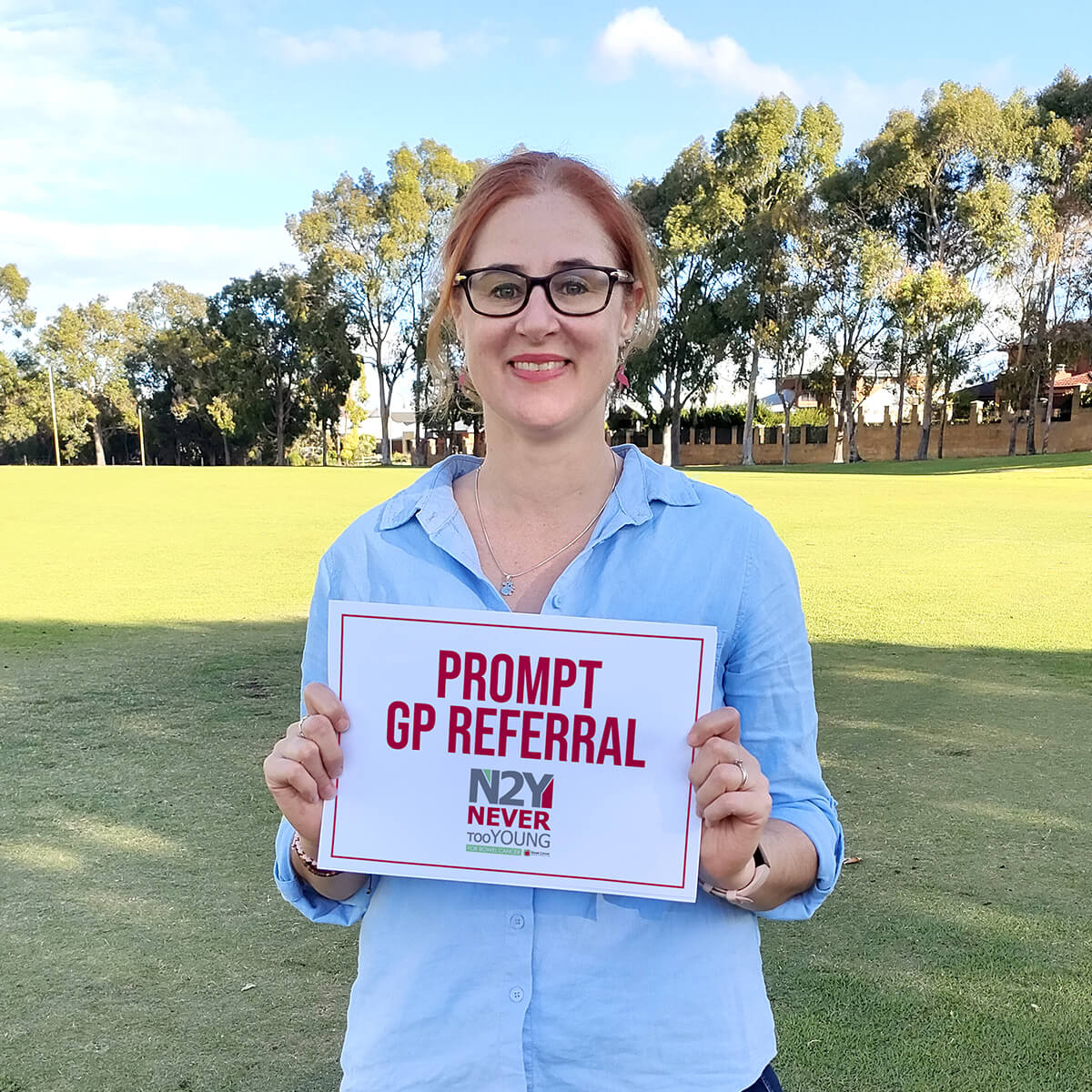 A woman with red hair and glasses holds a sign that reads "PROMPT GP REFERRAL" in a park setting.