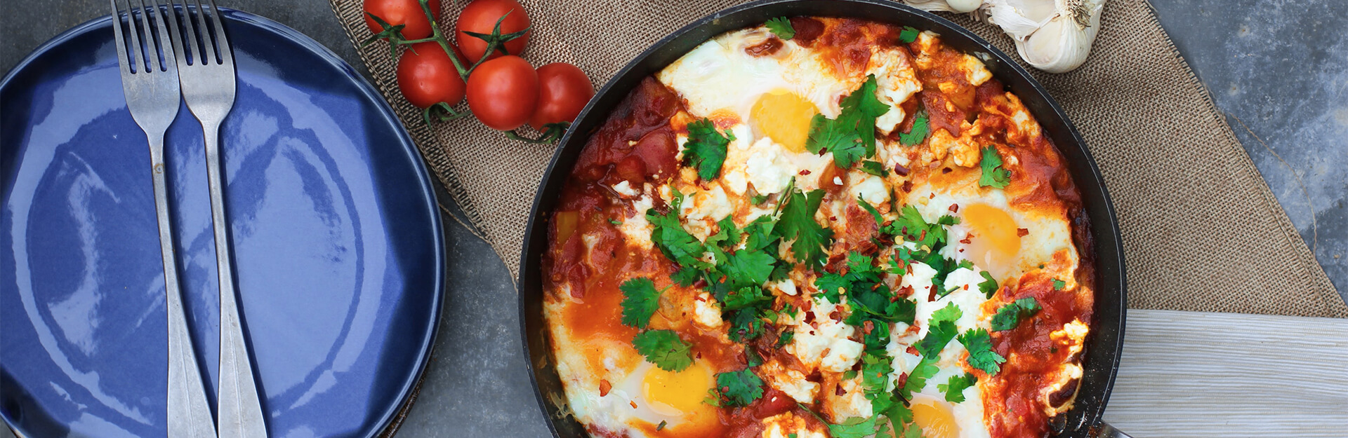 A skillet with eggs, tomatoes, and herbs, alongside a blue plate and forks, with cherry tomatoes and garlic nearby.