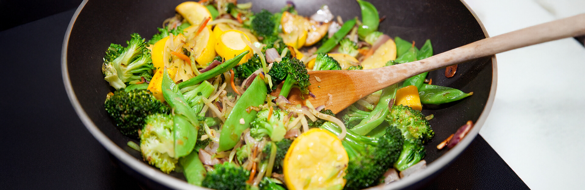 A skillet with sautéed broccoli, yellow squash, snap peas, and onions, with a wooden spoon stirring the vegetables.