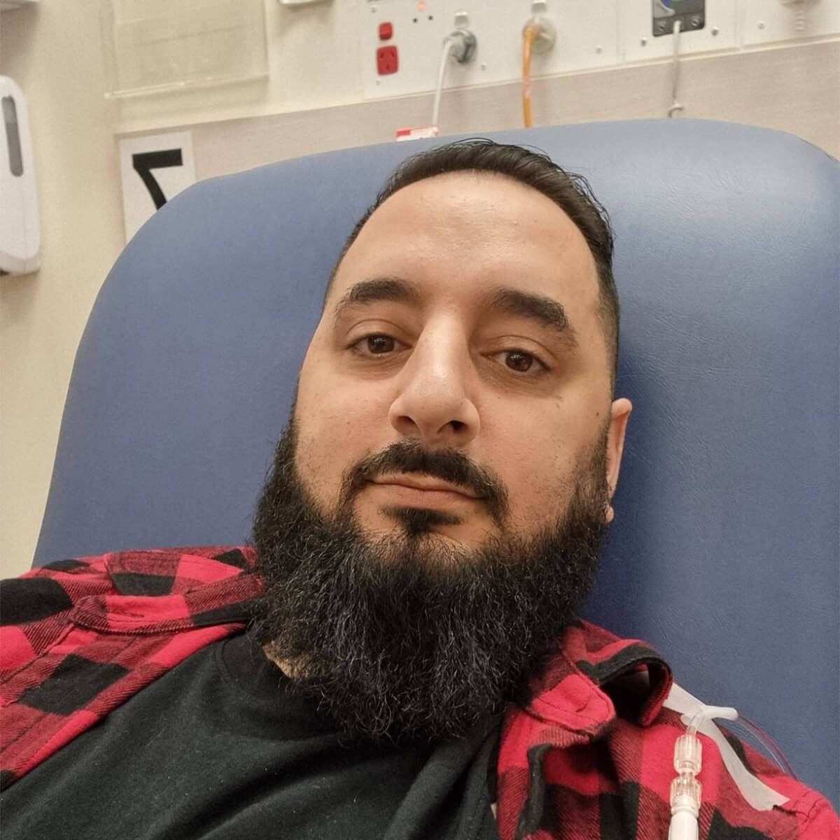 A man with a beard wearing a red and black flannel shirt is sitting on a hospital bed with an IV in his arm.