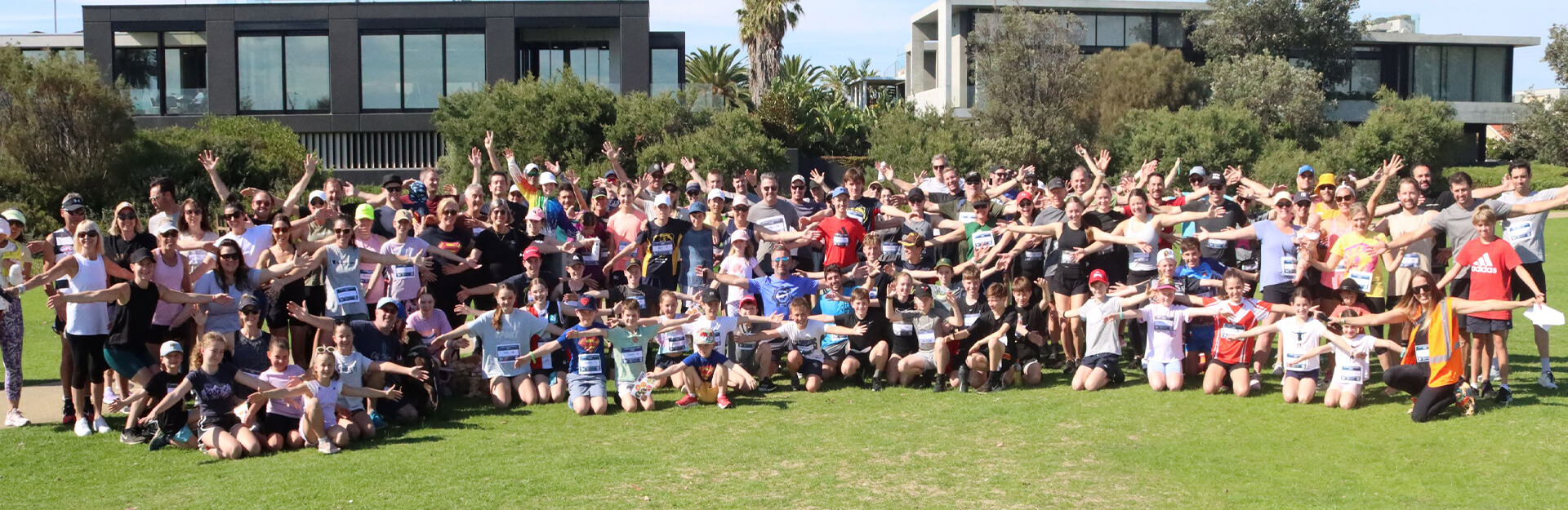 A large group of people poses outdoors, smiling and waving, with greenery and modern buildings in the background.