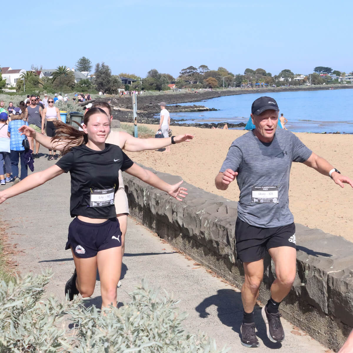 Two runners, a girl and a man, are jogging along a seaside path with spectators and a sandy beach in the background.