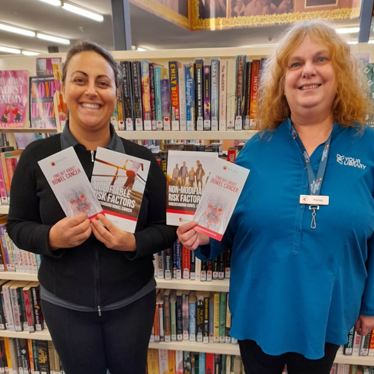 Two women hold pamphlets about bowel cancer in front of a bookshelf filled with various books.