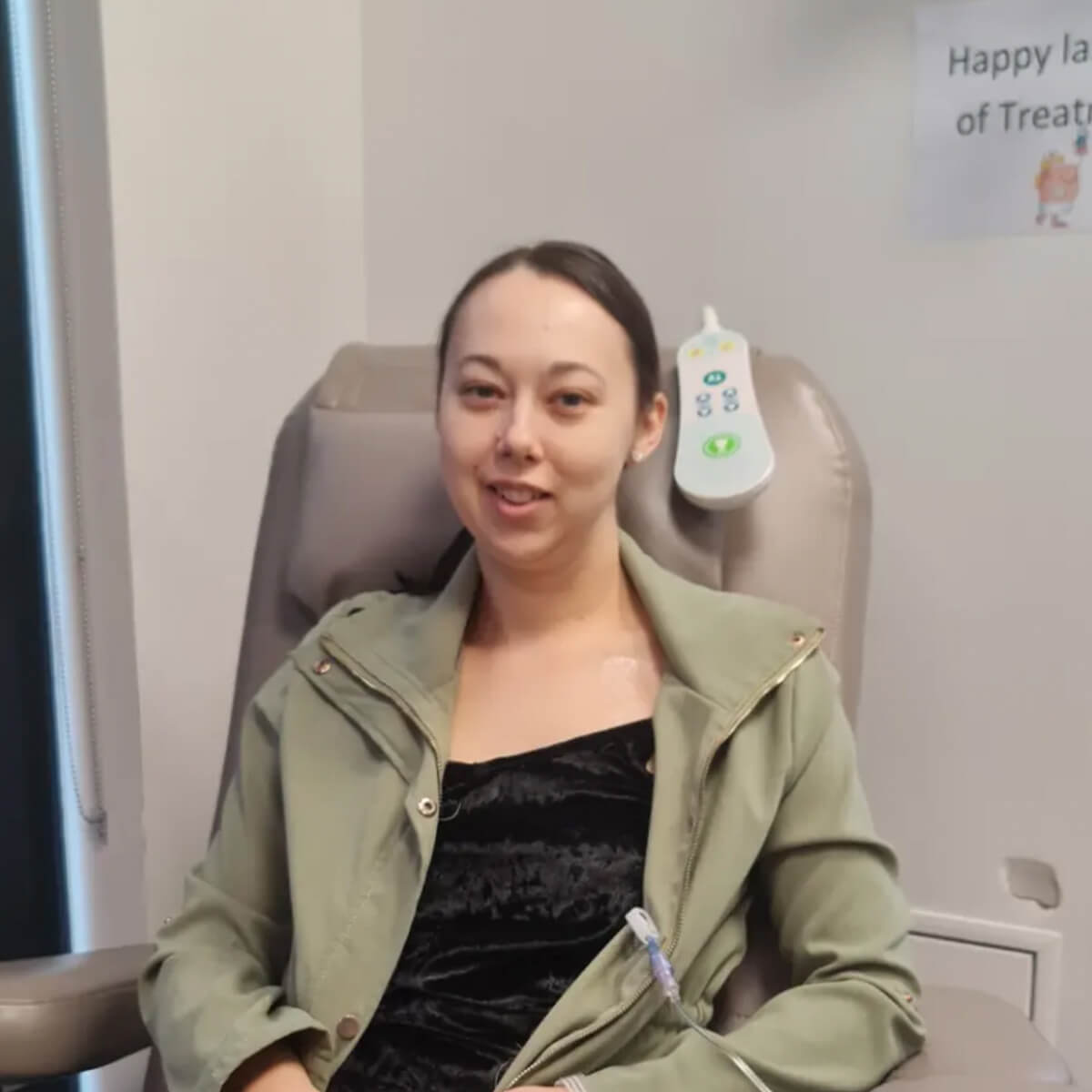 A woman sitting in a medical chair, wearing a green jacket, smiling, with an IV connected to her arm.