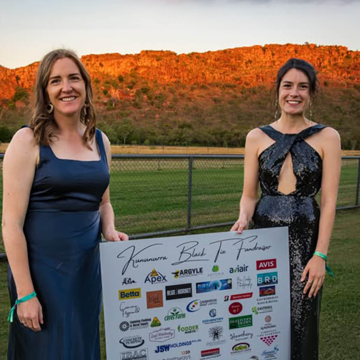 Two women in formal dresses hold a sign for a fundraising event, with a scenic background of hills at sunset.