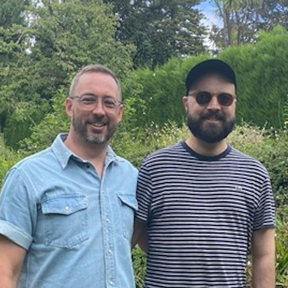 Two men stand together in a garden, smiling. One wears a light blue shirt and glasses, the other in a striped shirt and sunglasses.