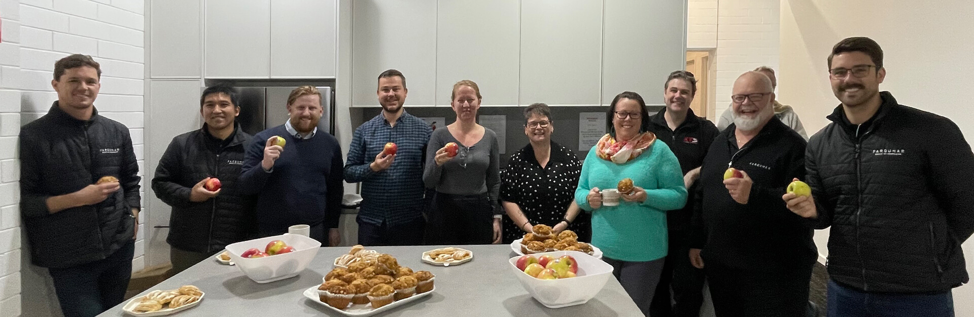 A group of nine people in a kitchen holding apples and pastries, smiling at the camera.
