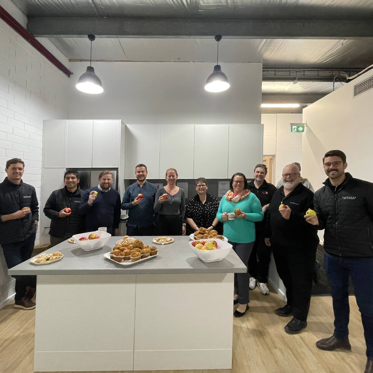 A group of ten people stands around a kitchen island with bowls of fruit and pastries, smiling and holding apples.