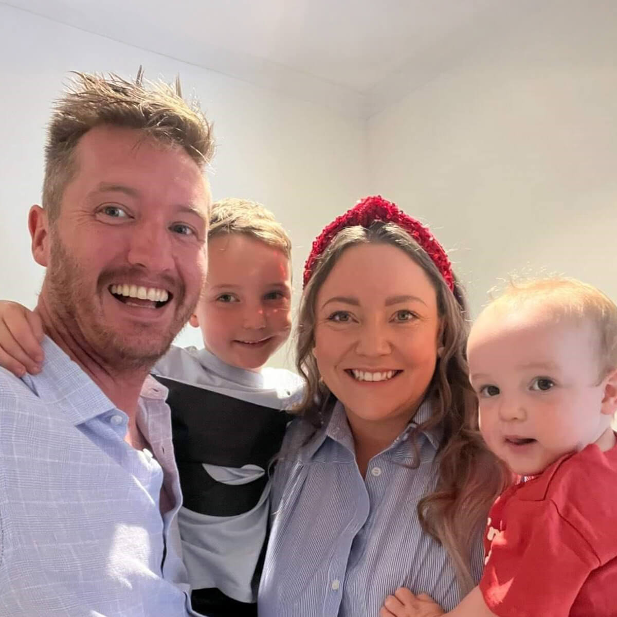 A smiling family of four poses together, with two children and two adults in a bright indoor setting.