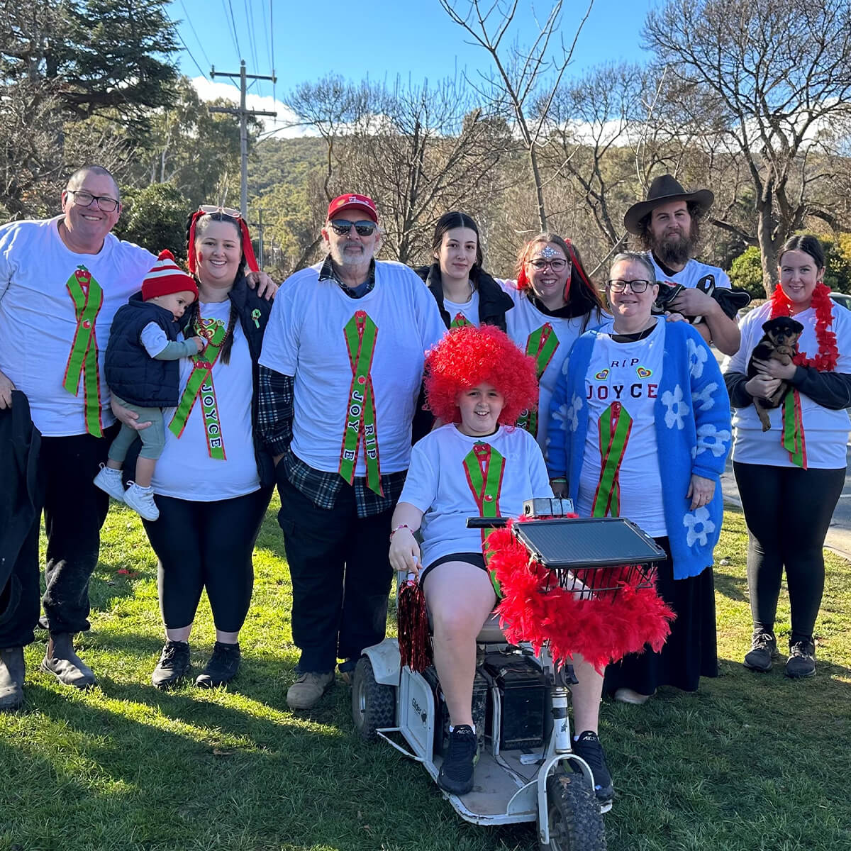 A group of nine people in matching shirts with festive designs, posing outdoors, some wearing red wigs and holding a dog.