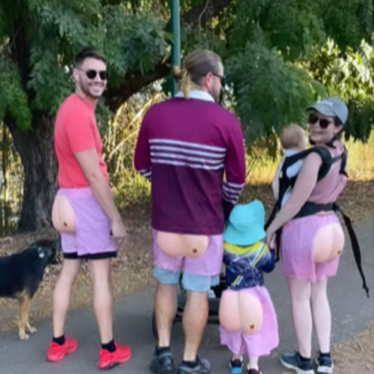 Four people wearing pink shorts with novelty butt designs stand outdoors, with a dog nearby and a child in a carrier.