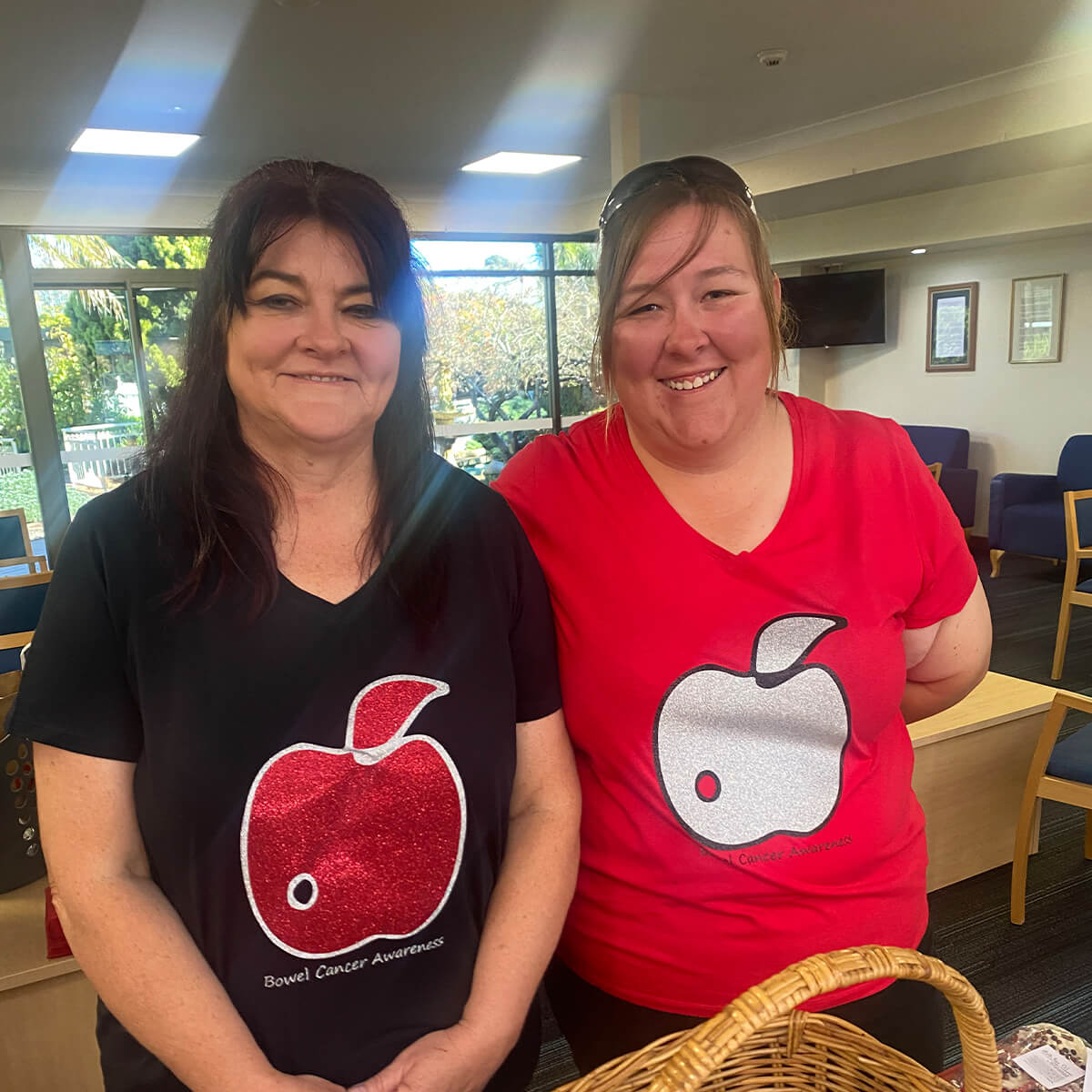 Two women stand side by side, wearing shirts promoting bowel cancer awareness, in a bright indoor setting.