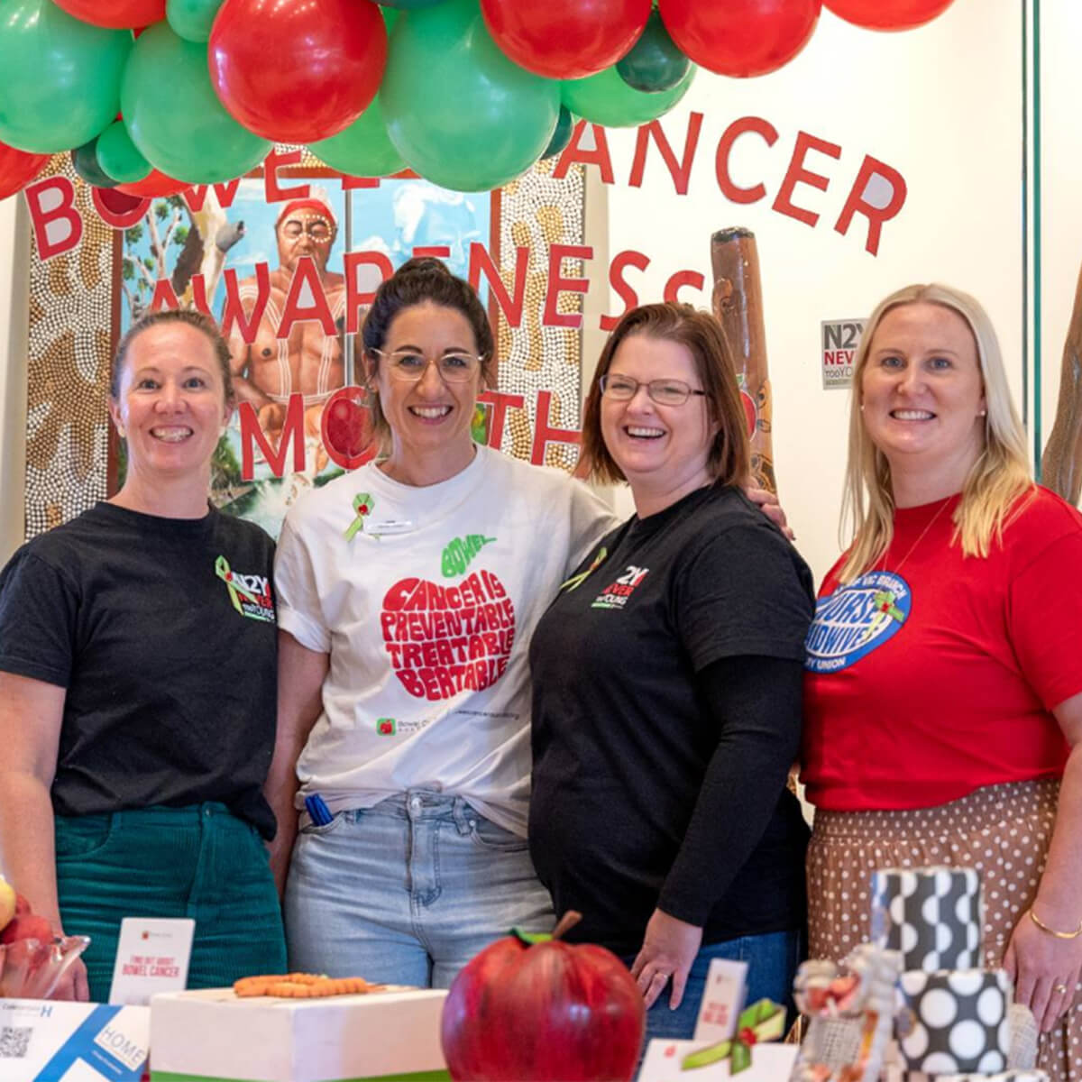 Four women stand together smiling at an event promoting bowel cancer awareness, with balloons and a display in the background.