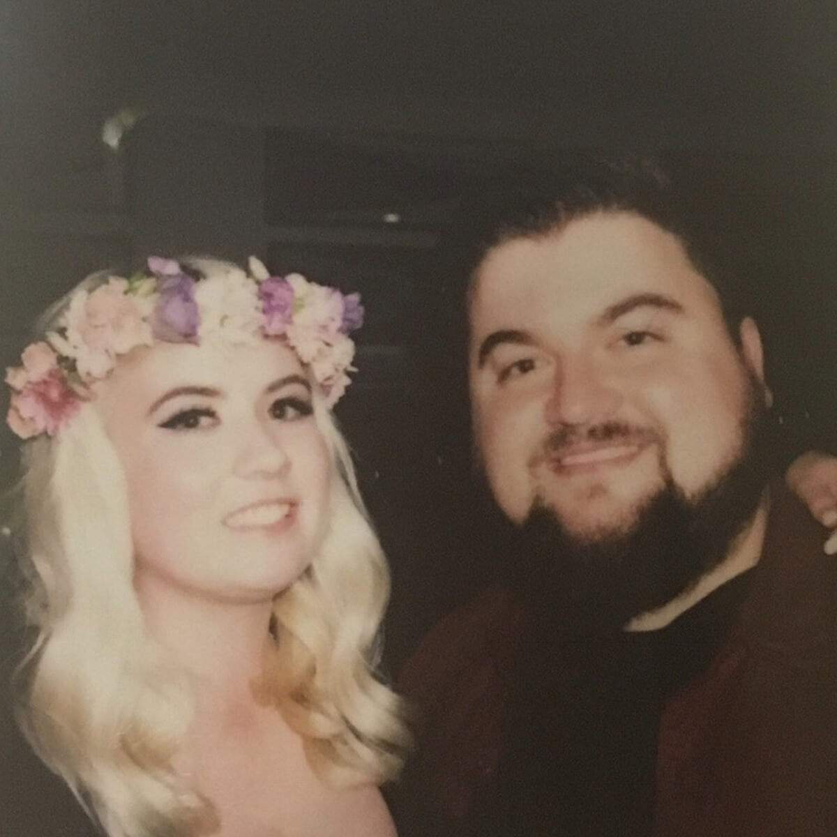 A smiling woman with blonde hair and a flower crown stands next to a bearded man, both in a dimly lit setting.