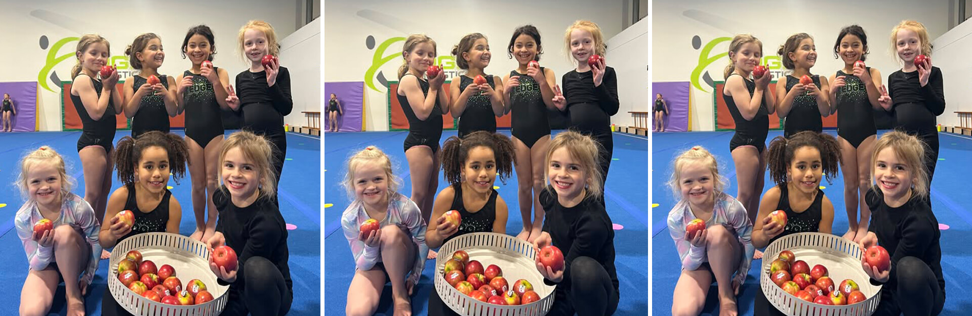 A group of young girls in gymnastics attire pose with apples in a gym setting, smiling and having fun.