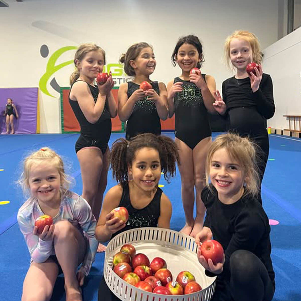 A group of eight young girls in gymnastics outfits holding apples, smiling, with a blue gym floor in the background.