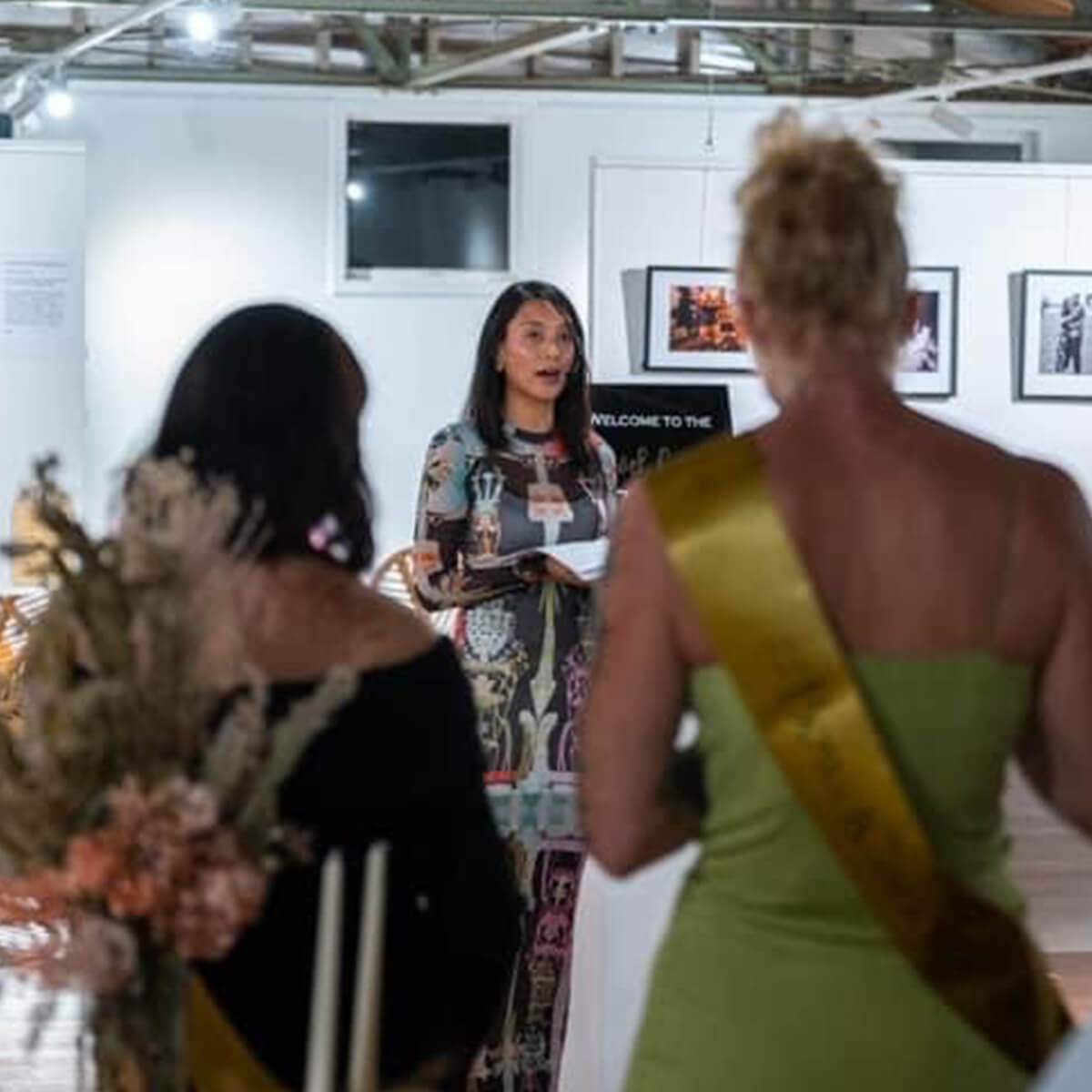 A woman in a patterned dress speaks at an event, with attendees listening and floral arrangements in the foreground.