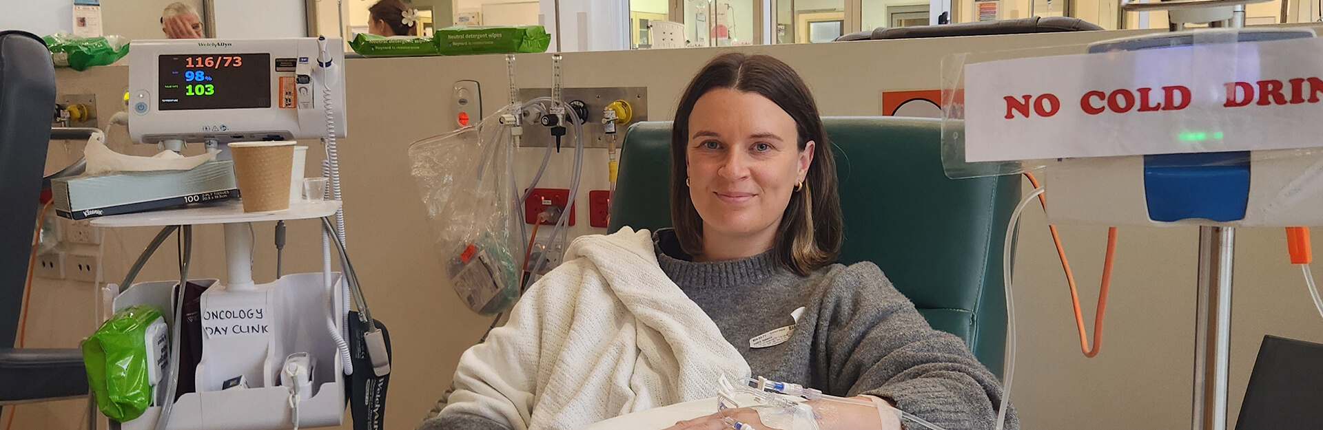 A woman sits in a hospital chair with an IV, smiling. Medical equipment and a sign reading "NO COLD DRINKS" are visible.