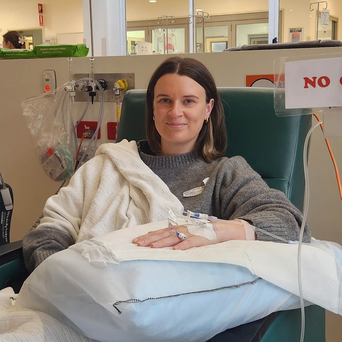 A woman sitting in a medical chair, wrapped in a blanket, with an IV in her arm and a calm expression.