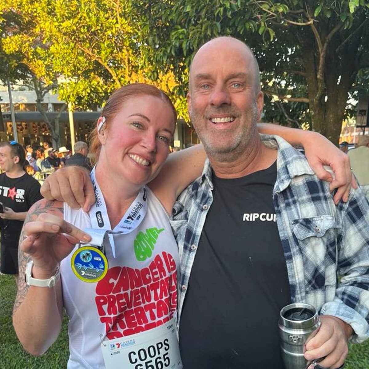 A woman in a race shirt holds a medal next to a smiling man, both outdoors with trees in the background.