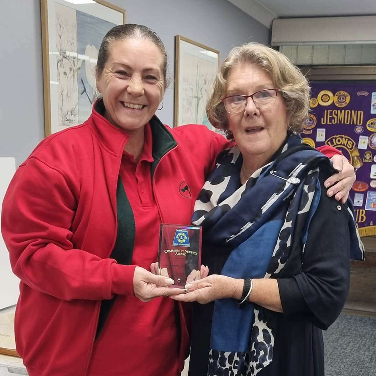 Two women smiling and holding a community service award, with a colorful backdrop of club banners.