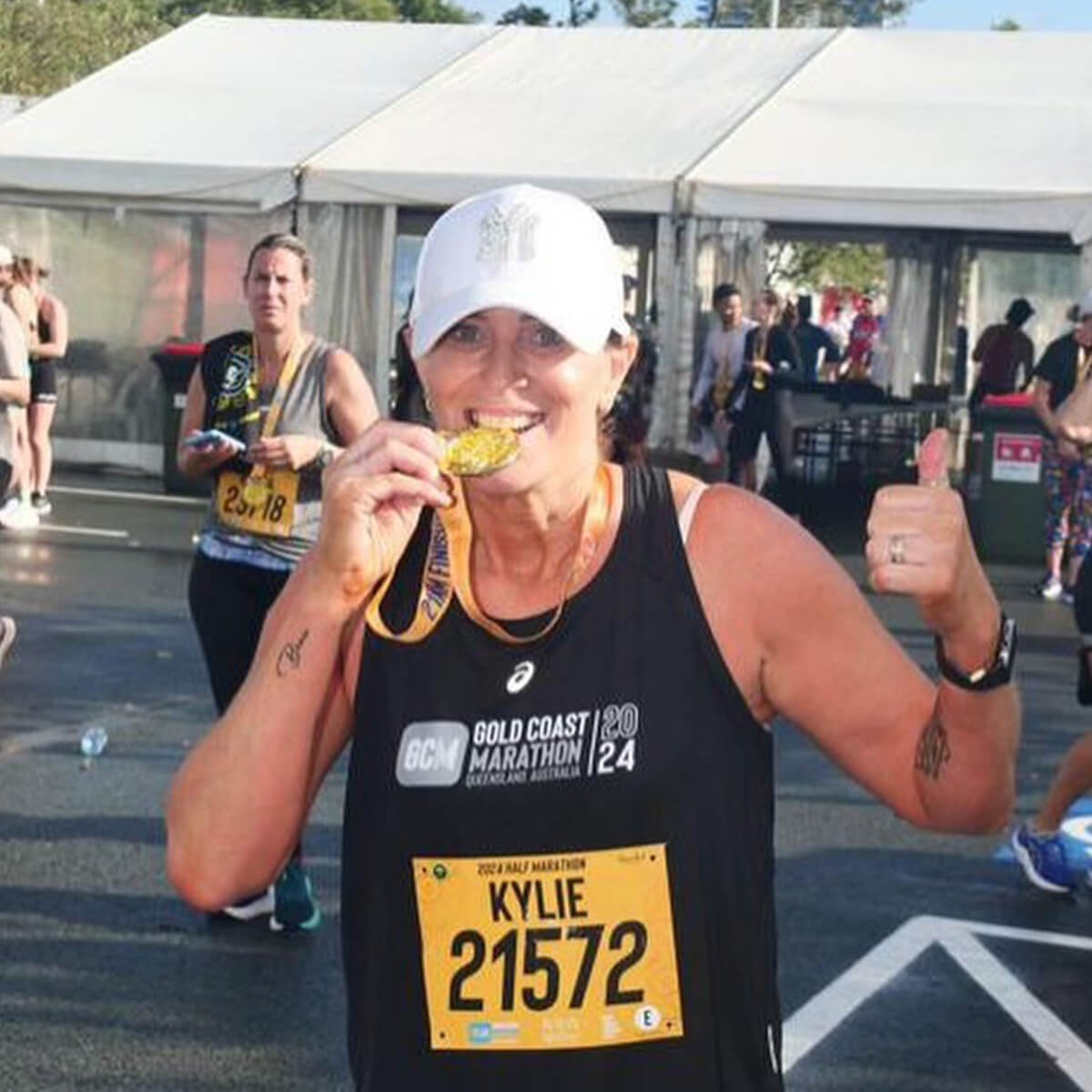 A woman in a marathon shirt smiles and holds a medal while giving a thumbs up, with other runners in the background.