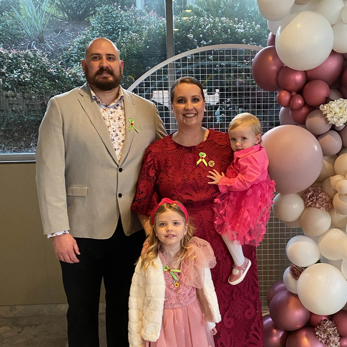 A family of four poses together, with two girls in pink dresses, a woman in a red dress, and a man in a light jacket.