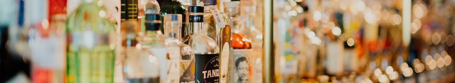 A blurred display of various liquor bottles on a bar shelf, with warm lighting creating a soft ambiance.