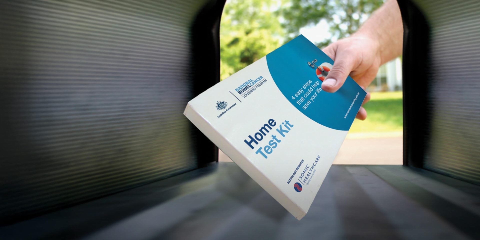 A hand is placing a "Home Test Kit" into a mailbox with a blurred background of green trees.