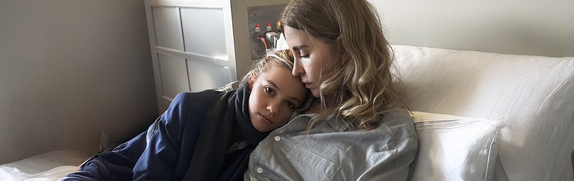 Two women sit closely together on a bed, one resting her head on the other's shoulder in a comforting pose.
