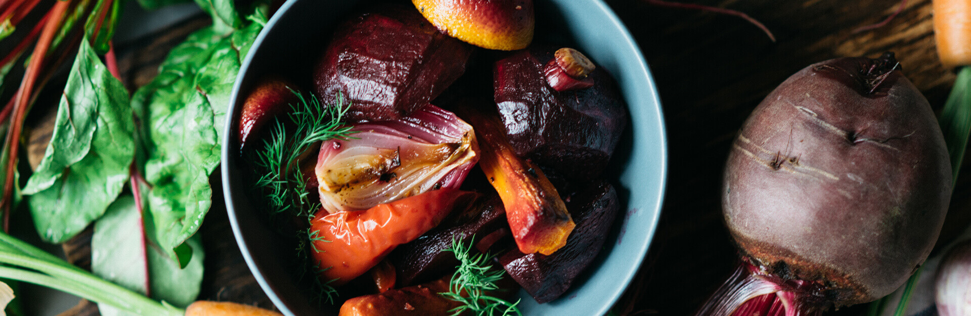 A bowl of roasted vegetables including beets, peppers, and onions, surrounded by fresh greens and carrots.