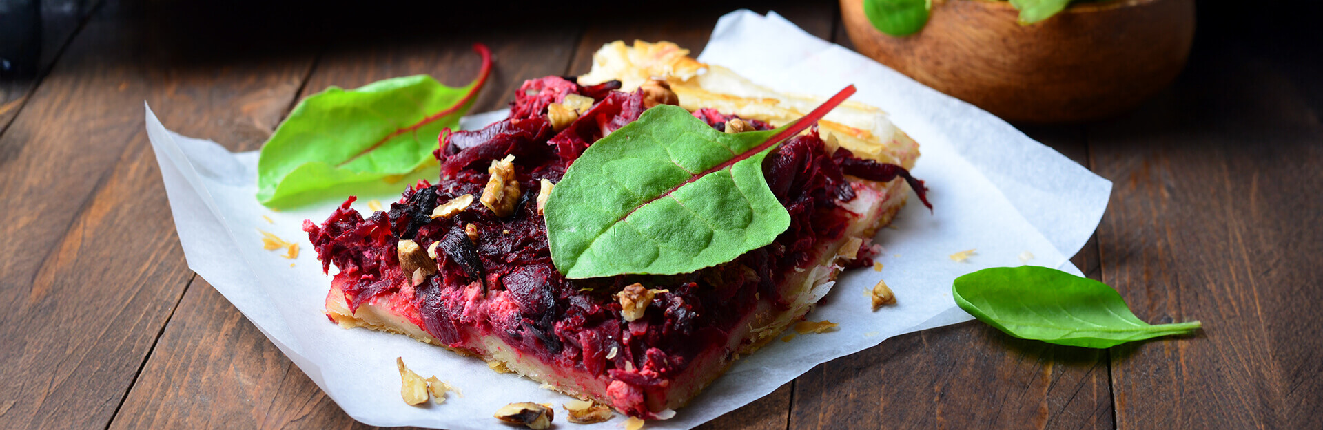 A slice of beetroot tart topped with spinach leaves and nuts, served on parchment paper on a wooden surface.