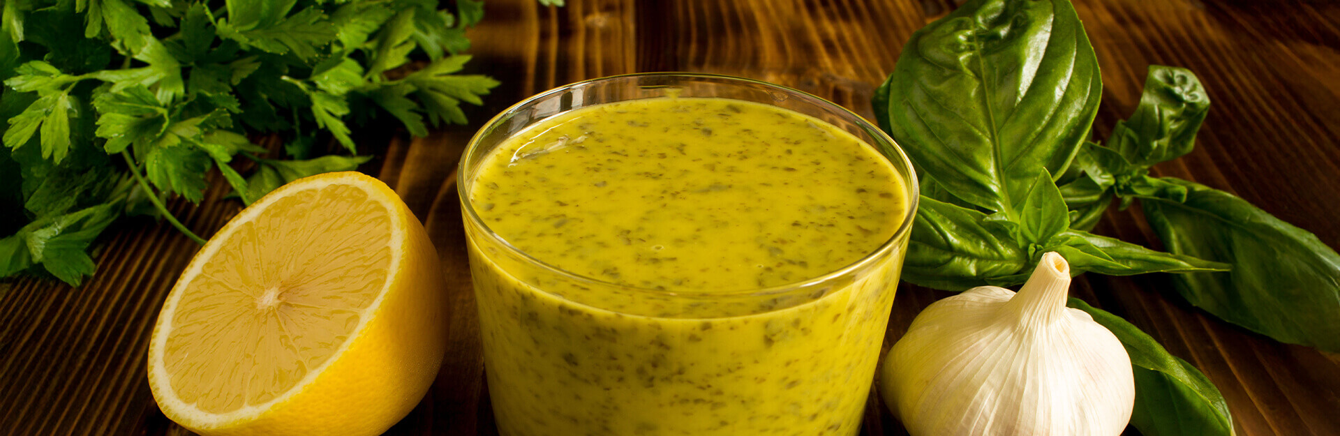 A glass of green sauce is surrounded by lemon, garlic, and fresh basil on a wooden surface.