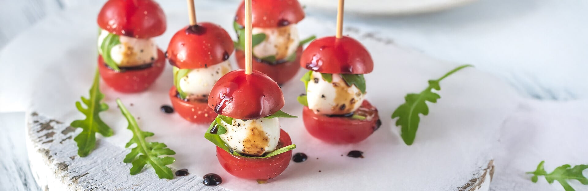 Five stacked tomato and mozzarella skewers with basil, drizzled with balsamic glaze, on a white plate.