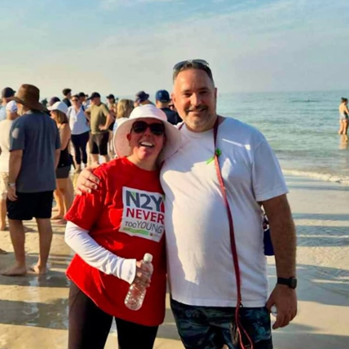 A woman in a red shirt and a man in a white shirt pose together on a beach with a crowd in the background.