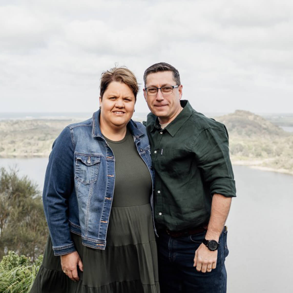 A woman in a denim jacket and a man in a green shirt stand together with a scenic view of a lake and hills behind them.
