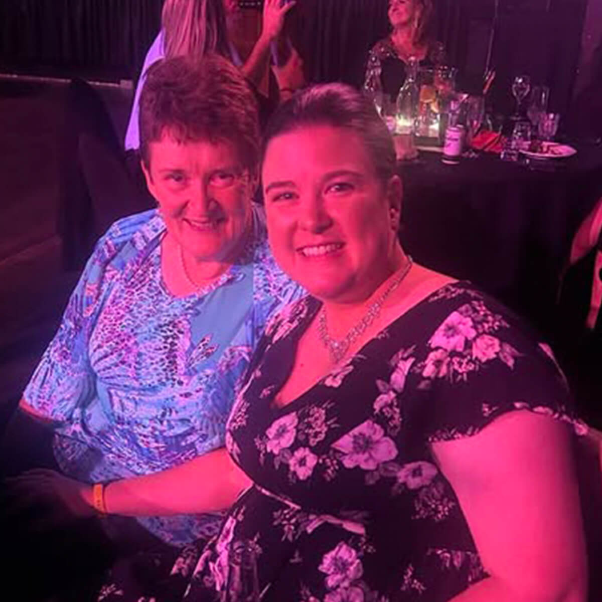 Two women sitting together, smiling at the camera, in a dimly lit venue with blurred background elements.