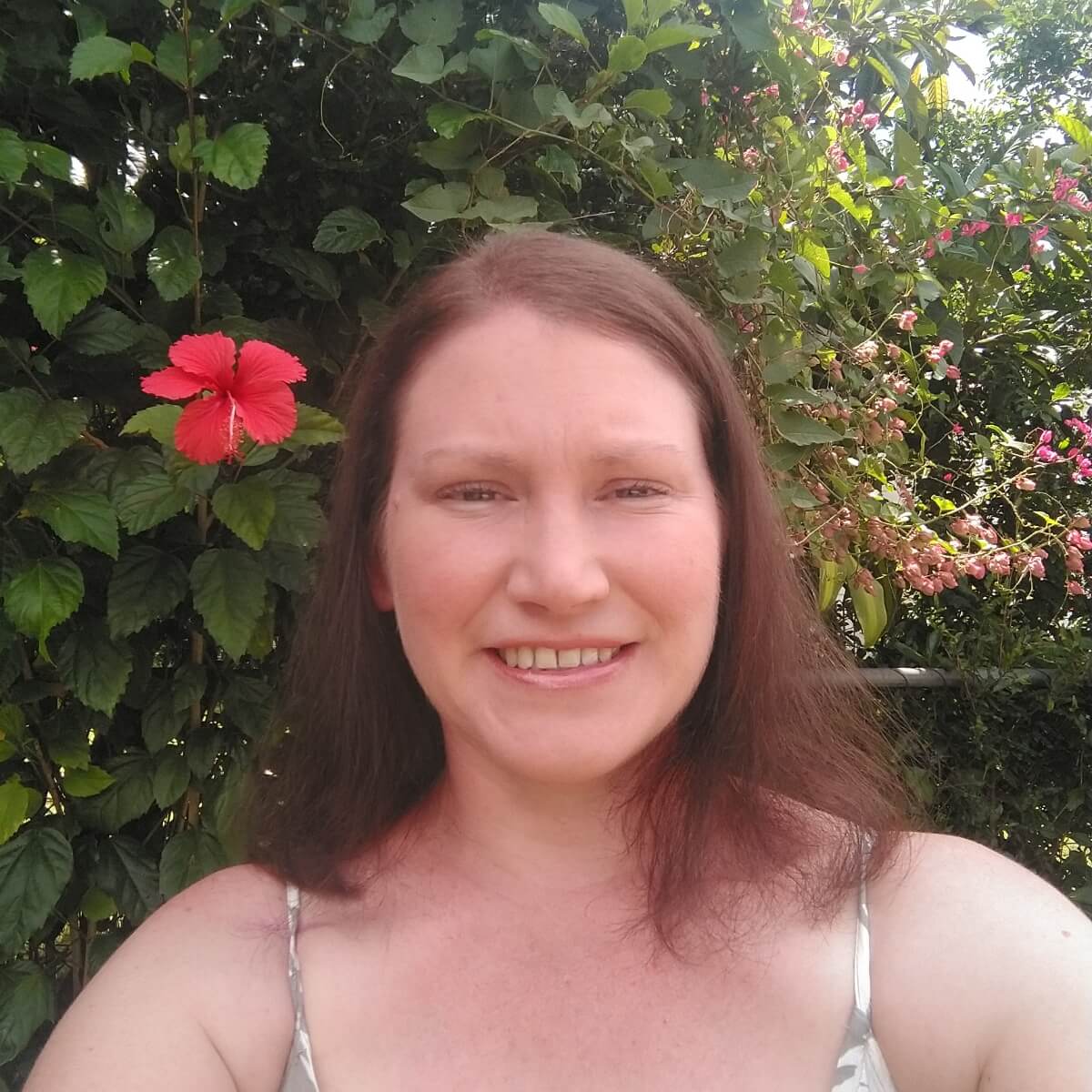 A woman with long brown hair smiles in front of lush greenery and colorful flowers.