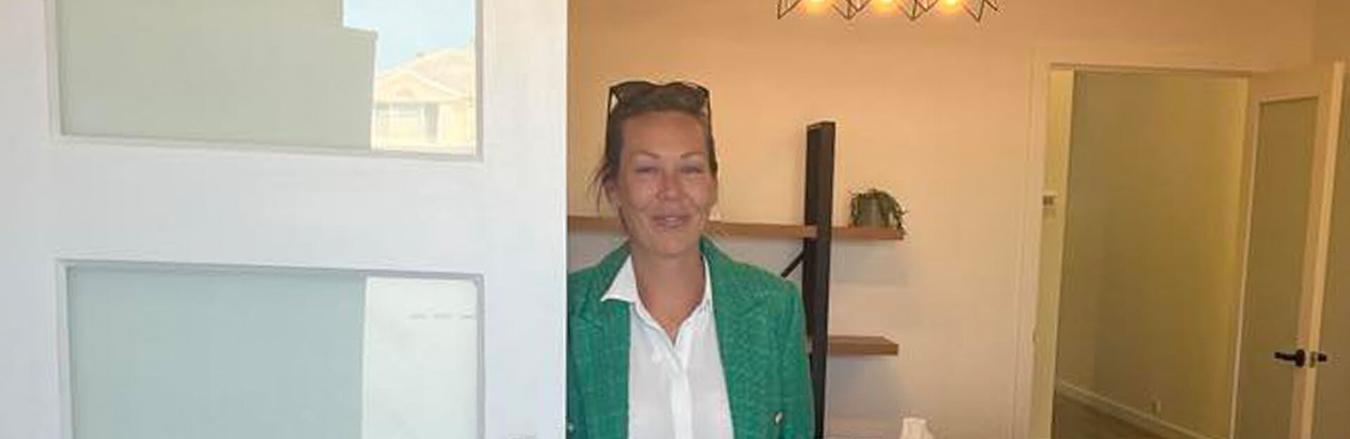A woman in a green blazer stands at a doorway, smiling, with a bright interior behind her.