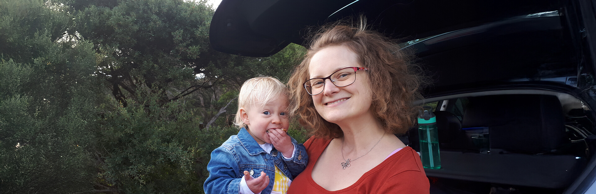 A smiling woman holds a toddler near a car, surrounded by trees and greenery.