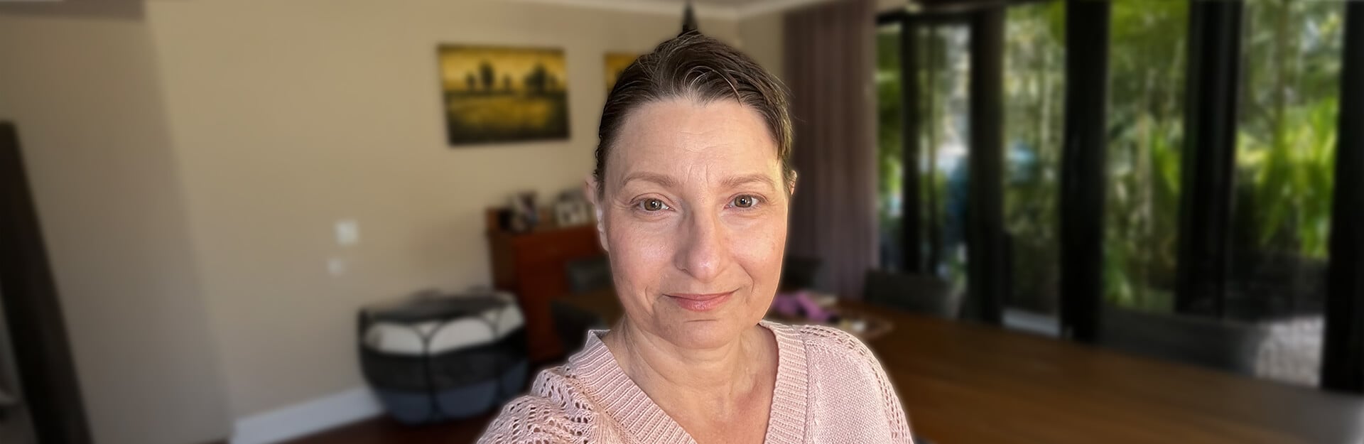 A woman with short brown hair and a light pink sweater stands in a bright room with large windows and greenery outside.