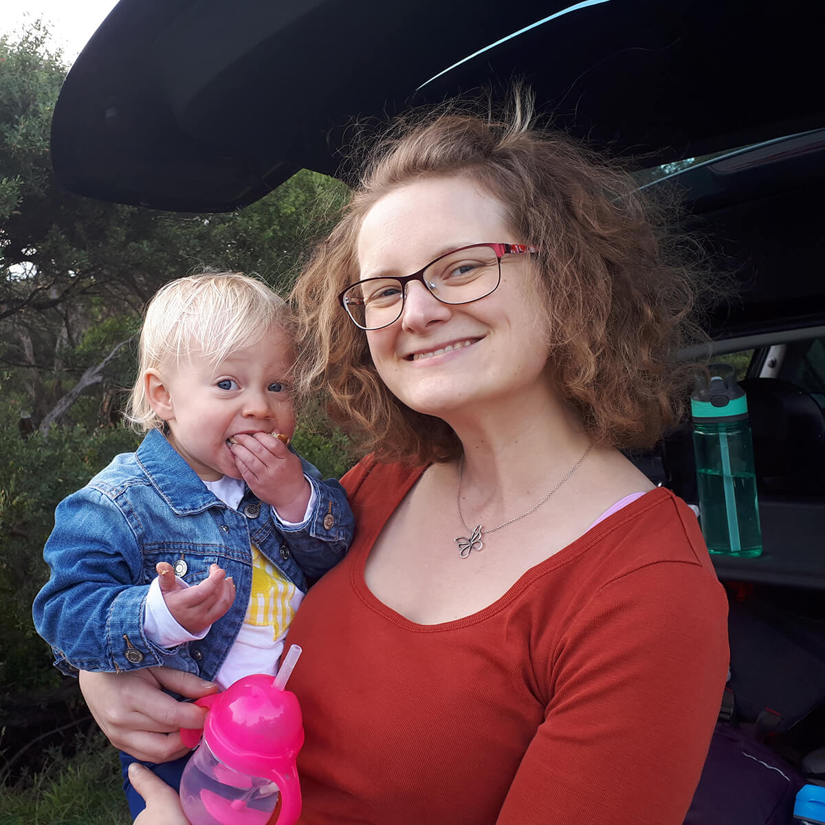 A woman with curly hair and glasses smiles while holding a toddler eating a snack, near a car in a natural setting.