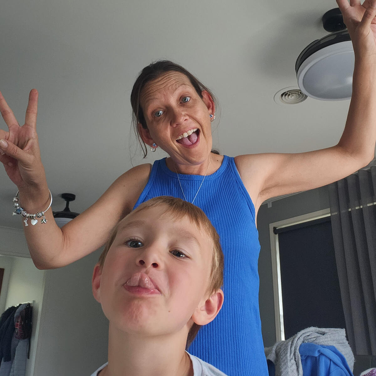 A woman in a blue dress poses playfully with a child making a silly face, both smiling in a bright room.