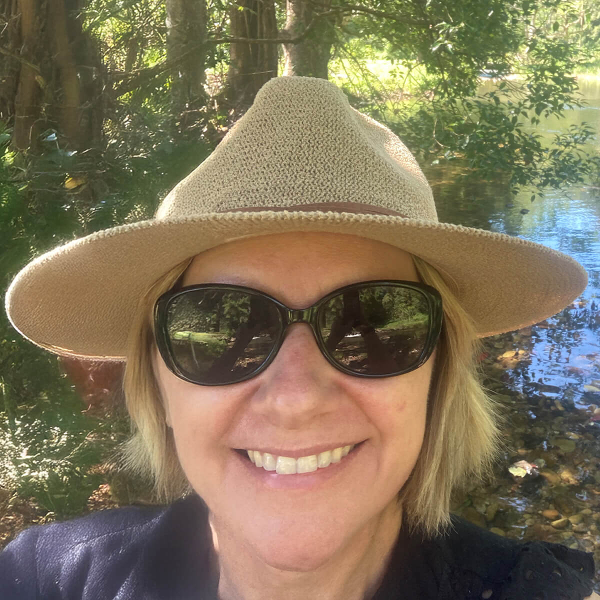A woman with short blonde hair wearing a wide-brimmed hat and sunglasses smiles by a tranquil river surrounded by greenery.